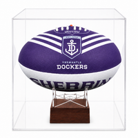 A purple and white Fremantle Dockers rugby ball displayed inside a clear acrylic cube case on a dark wood stand.