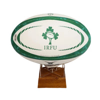 A white rugby ball with green trim and a green shamrock logo with the text 'IRFU' on the front, displayed on a small wooden stand against a plain white background.