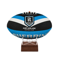 A custom Port Adelaide Football Club rugby ball urn with a black, blue, and white striped design, displayed on a clear acrylic stand with a dark brown wooden base against a white background.