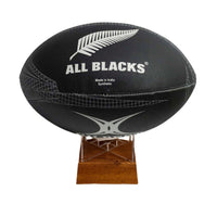 A black All Blacks rugby ball with silver fern and white branding, displayed on a small wooden stand against a plain white background.