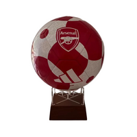 A red and white patterned Arsenal soccer ball mounted on a clear acrylic stand with a dark wooden plaque base, set against a plain indoor wall.