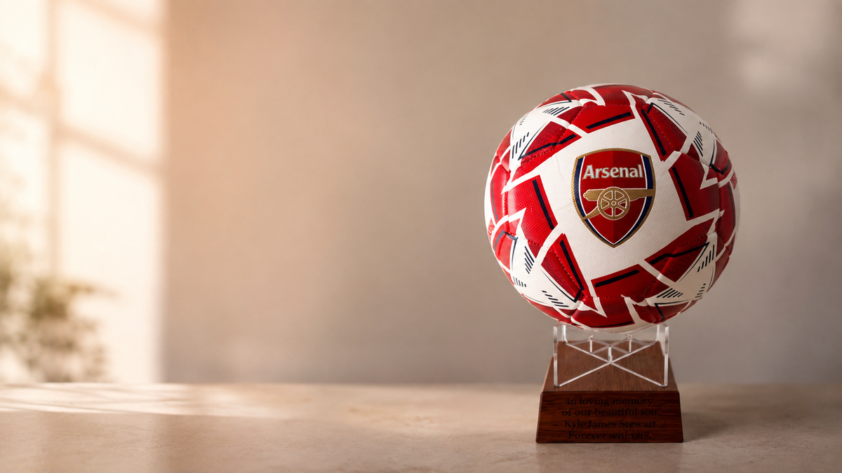 An Arsenal soccer ball with a red and white pattern, displayed on a clear acrylic stand atop a wooden plaque, set against a warm, softly lit indoor background.
