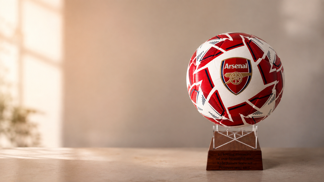 An Arsenal soccer ball with a red and white pattern, displayed on a clear acrylic stand atop a wooden plaque, set against a warm, softly lit indoor background.