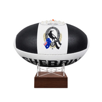 A black and white AFL ball with the Collingwood Football Club logo, displayed on a clear stand with a dark brown wooden base against a white background.