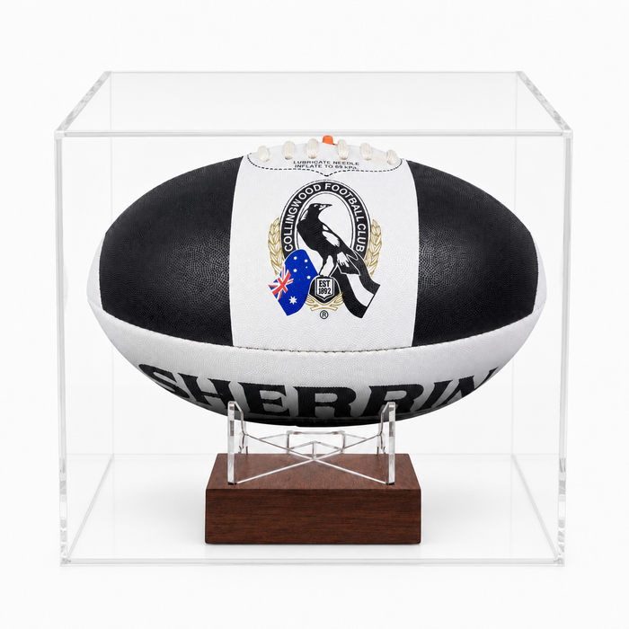 A black and white AFL ball with a Magpies logo, displayed inside a clear acrylic case on a wooden stand.