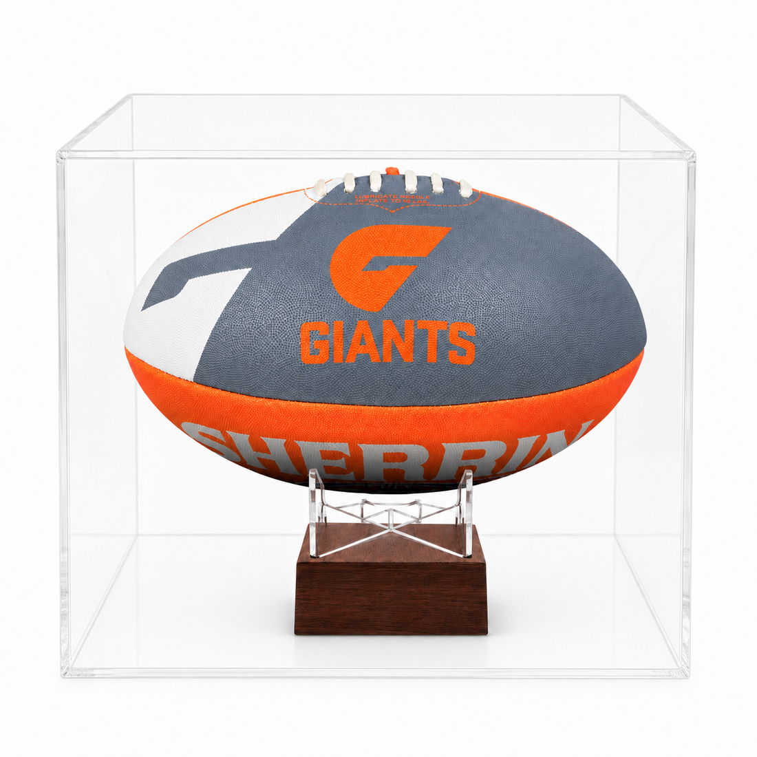 A Sherrin football displayed inside a clear acrylic display case on a small wooden stand. The ball is gray, orange, and white with the text &