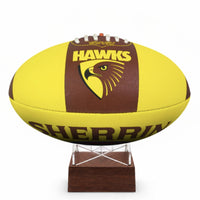 A yellow and brown Sherrin Hawks football displayed on a small wooden stand against a white background.