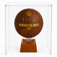 A vintage brown leather Manchester United soccer ball urn with gold lettering and crest, displayed inside a clear acrylic shadowbox case on a wooden stand.