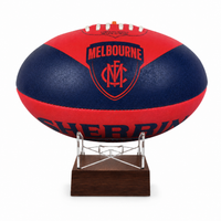 A red and navy blue Melbourne Football Club AFL ball urn displayed on a clear acrylic stand with a dark wood base against a white background.