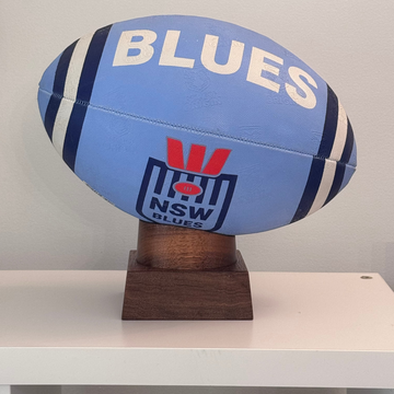 Blue and white rugby ball with 'NSW Blues' branding on a wooden stand against a gray background.