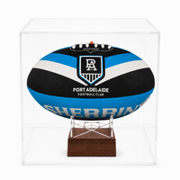 A Port Adelaide Football Club rugby ball displayed inside a clear acrylic cube case on a wooden stand.