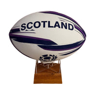 A white Scotland rugby ball with purple and navy blue stripes and the word 'SCOTLAND' printed on it, displayed on a small wooden stand against a plain white background.