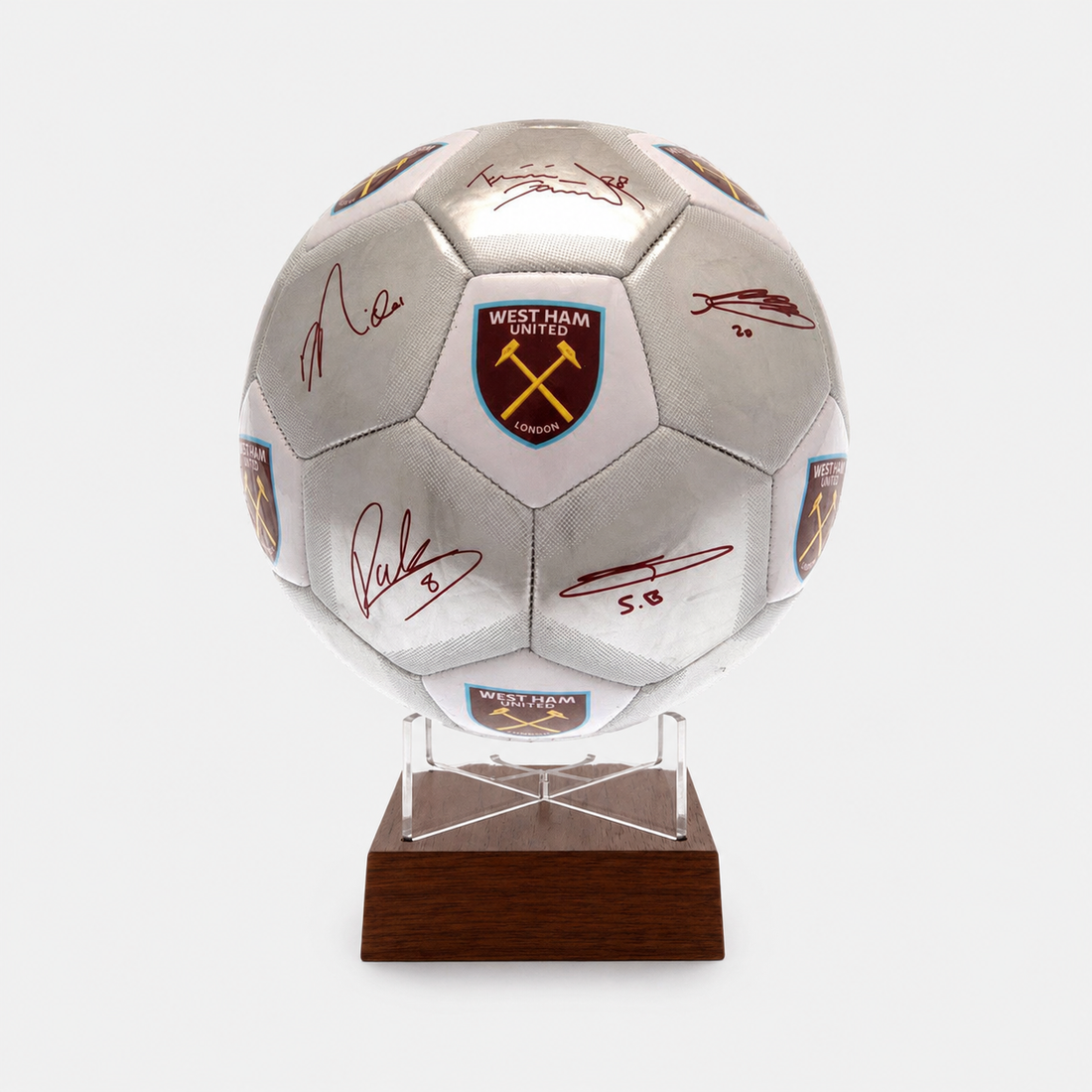 A silver West Ham United soccer ball with multiple autographs and team crests, mounted on a clear acrylic stand with a dark brown wooden base against a light gray background.