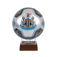 A silver and black soccer ball featuring the Newcastle United crest and multiple player signatures, displayed on a clear stand with a dark wood base against a light gray background.